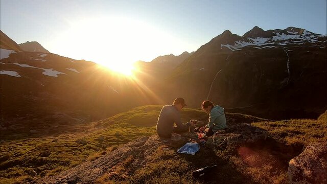 A couple camping in the Austrian Alps near Schladming. They are preparing their meal with the sun setting behind the high mountain peaks. Some snow in the higher parts. Camping in the wilderness. Free