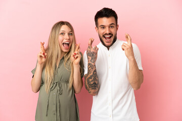 Pregnant woman and man over isolated pink background with fingers crossing