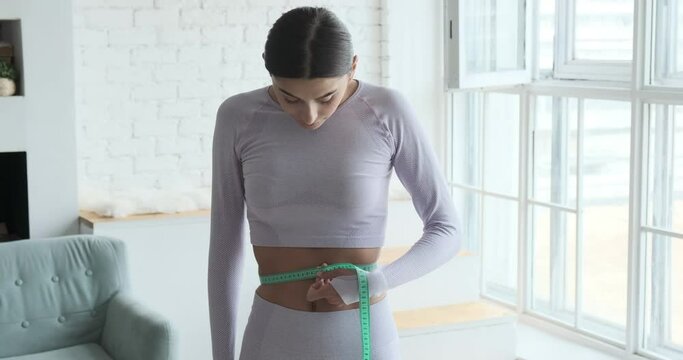 Athletic lady measures bare waist with tape standing at home after fitness exercises workout close view. Healthy diet lifestyle and fat burn slimming weight loss concept.