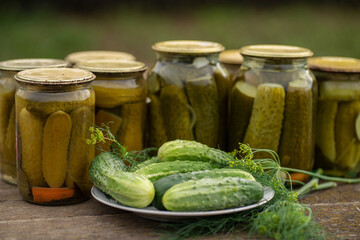 Glass jars with canned cucumbers in a rustic style on a wooden table. Plate with fresh cucumbers and dill