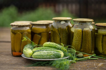 Glass jars with canned cucumbers in a rustic style on a wooden table. Plate with fresh cucumbers and dill