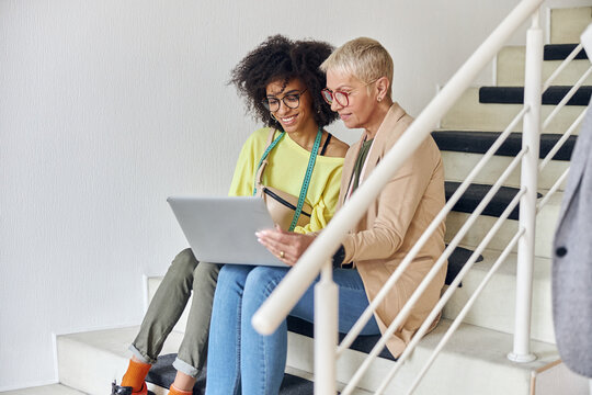 Mature woman and young African-American use laptop stitting on staircase in design studio
