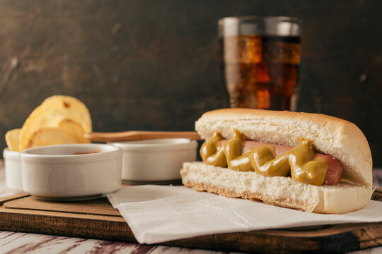Normal view of a hotdog in the foreground with a glass of cola behind it and some potato chips. Fast food concept.