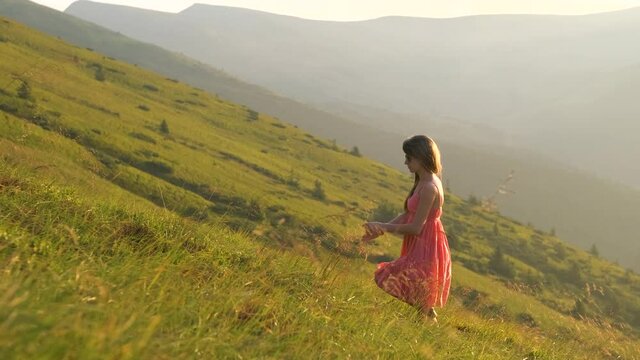 Young woman in red dress walking on hillside meadow on a windy evening in summer mountains.