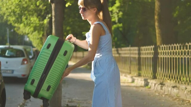Young female tourist taking out green suitcase from car roof rack and walking on city street. Travel and vacation concept.