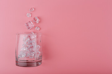 Creative flat lay with empty transparent glass with spilled ice cubes on pink background with copy space. Concept of summer hot, refreshing iced summer drinks and cold cocktails