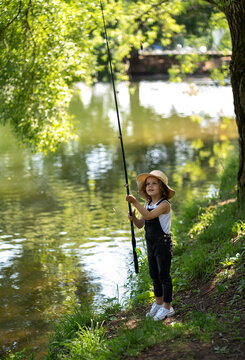 A little girl with curly hair in a straw hat and black overalls stands with a fishing rod by the pond. Child fishing on a summer day