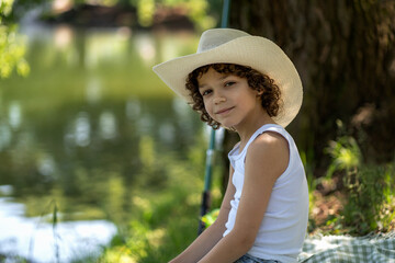 Portrait of a cute boy with curly hair in a straw hat by the pond. Child fishing on a summer day