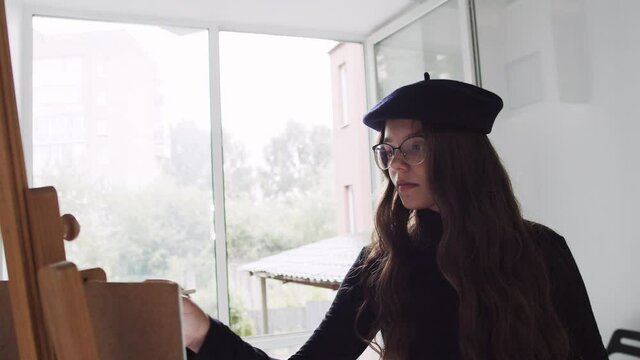 Young woman artist in beret painting picture on canvas in art studio