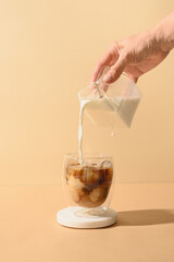 Pouring milk in coffee with ice cube on modern beige background. Vertical format. Iced latte coffee...