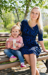 A young beautiful pregnant woman and a little toddler daughter are sitting on park bench. A sunny summer day