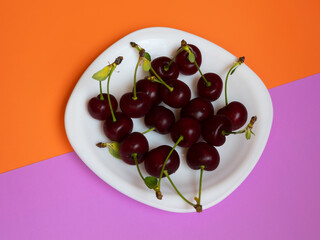 A serving of red cherries on a white plate lies on a double background, beveled towards a purple-orange background.