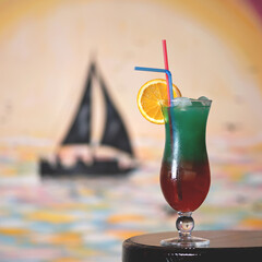 Iced summer cocktail. Cold drink on table. Two-layer red-green beverage in glass with orange slice and straw. Sailboat on blurred background. Copy space. Soft focus.