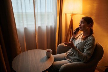 Obraz na płótnie Canvas Communication. A woman is sitting in a chair in the living room and talking on the phone. There is a cup of coffee on the table in front of her. There is a floor lamp in the corner of the room.