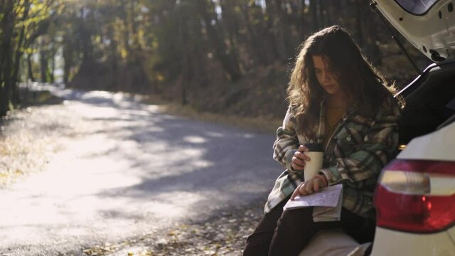Curly haired young woman in checkered jacket drinks tea and reads paper map sitting in open trunk of hatchback car on empty rural road in autumn forest