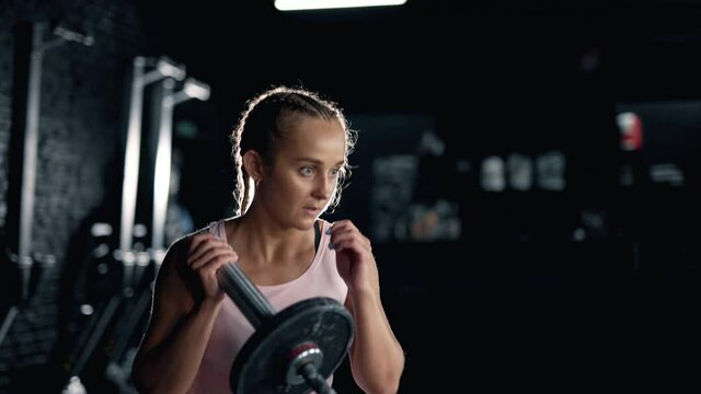 A muscular sportswoman is doing exercises in the dark gym. Self confident girl is engaging with barbell. Crossfit training. Maximum focus and strength. Concept of lifestyle fitness health and sports.