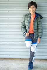 Full length Portrait of young boy looking towards the camera outdoors in natural light standing against corrugated wall..
