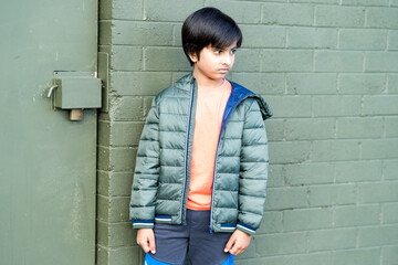 Portrait of young boy looking away from the camera outdoors in natural light. Male kid wearing puffer jacket standing against green brick wall and door.
