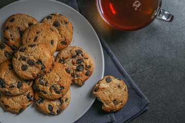 A cup of hot black tea in a glass cup and a gray ceramic plate with cookies with chocolate chips on a gray ceramic counter top with a gray linen napkin.