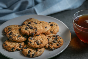 A cup of hot black tea in a glass cup and a gray ceramic plate with cookies with chocolate chips on a gray ceramic counter top with a kitchen towel.