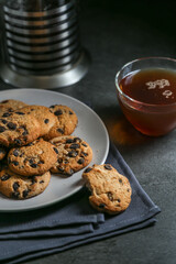 A cup of hot black tea in a glass cup and a teapot press and a gray ceramic plate with cookies with chocolate chips on a gray ceramic counter top with a gray linen napkin.