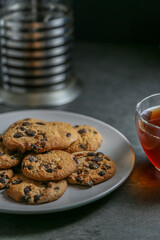 A cup of hot black tea in a glass cup and a teapot press and a gray ceramic plate with cookies with chocolate chips on a gray ceramic tabletop.
