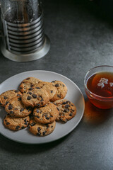 A cup of hot black tea in a glass cup and a teapot press and a gray ceramic plate with cookies with chocolate chips on a gray ceramic tabletop.