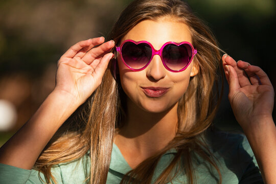 Portrait of young woman standing outside in bright sun wearing heart shaped sunglasses, looking into camera lens 