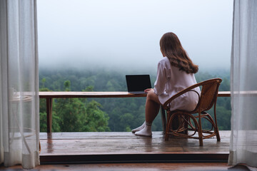 Rear view image of a woman using and working on laptop computer while sitting on balcony with a beautiful nature view on foggy day