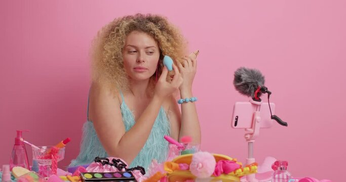 Attractive curly haired woman applies foundation on face with sponge gives tutorial for followers how to do makeup wears blue dress sits near table full of cosmetic products isolated on pink wall