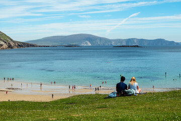 Fototapeta na wymiar Beautiful couple sitting on a grass and enjoy great view on the ocean. Keem bay and beach, Achill island, Ireland. Warm sunny day. Love and romance concept. Travel and outdoor activity