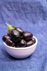 Closeup of Organic Malabar Plum or Jamun in a Bowl Isolated on Blue Fabric Background with Leaf in Vertical Orientation, Also Known as Syzygium Cumini, Java Plum or Jambolan