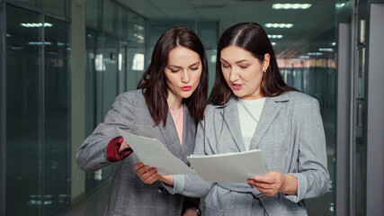 Obraz premium Successful women company managers in grey jackets discuss report holding papers in hands standing in company building corridor closeup