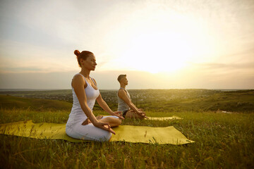 Woman and man sitting in lotus position with eyes closed, meditating and practicing yoga together on nature outdoors at sunset or sunrise on summer day. Yoga practice, healthy lifestyle concept