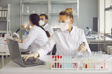 Scientists doing research in well-equipped medical science laboratory. Lab worker or assistant in protective face mask and glasses entering data analysis report in electronic system on laptop computer