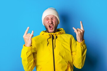 A young man in a yellow jacket with closed eyes shows a rocker goat, a horn on a blue background