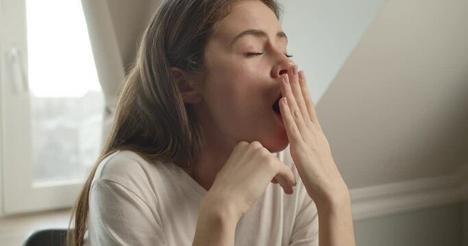 Tired girl brunette of Slavic appearance sits in a white T-shirt at the desk, yawns, looking aside and covering her mouth with her hand, wants to sleep against the window in the room.