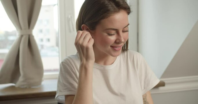 A cute brunette girl of Slavic appearance sits in a white T-shirt at the desk, on which there is a laptop, takes an earpiece, sticking it in her ear, listening to music in headphones and dancing