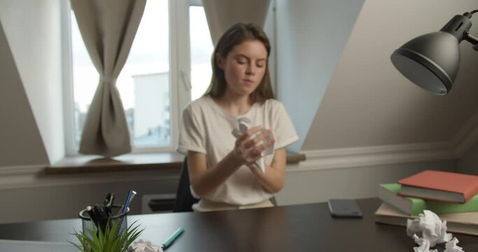 A frustrated brunette girl of Slavic appearance sits in a white T-shirt at the desk, on which there is a phone, notebooks, pens and a lot of wrinkled paper, wrinkles sheets of paper
