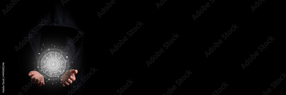 Wall mural young woman's face is not visible in a black hood holding with palms a glowing zodiac circle on a bl