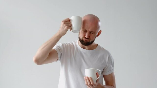 Morning hangover. Headache relief. Unhealthy lifestyle. Sick unwell suffering man drinking coffee water fast applying cold mug to head isolated on light neutral background.