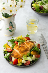 Delicious hearty lunch, chicken breast baked with spices and a mix of salad with tomatoes, radishes and cauliflower, a plate of salad and daffodils in a vase