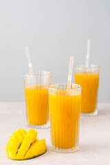 Freshly squeezed mango juice in glasses with a straw, ripe cut mango on a gray background