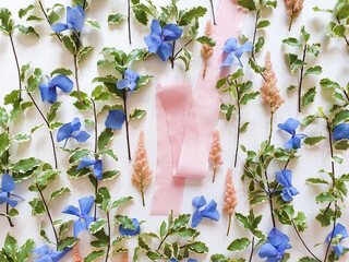 Pink silk ribbon, fresh greens, blue and pink flowers on white background. Cosmetics product advertising backdrop or background. Empty place to display product packaging. Showcase mockup, template.