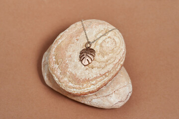 Gold metal chain and pendant in the shape of a monstera leaf on rocks over beige background