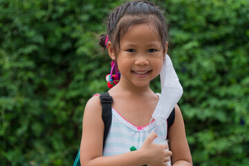 Fototapeta premium Little asian girl wearing face mask with green back pack holding stationery in green nature park background. Back to school concept.