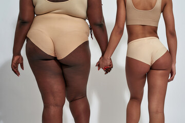 Cropped shot of two african american women in underwear with different body size holding each other hands, standing isolated over gray background