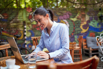  young beautiful caucasian adult female sitting in outdoor cafe, smiling, looking at her laptop, typing. online communication concept