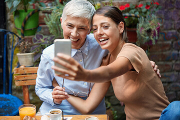 young adult caucasian female and elderly woman smiling, looking at cell phone,  taking selfie,  smiling, sitting in outdoor cafe.