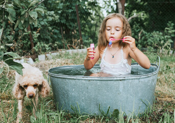 little cute girl in a pink swimsuit blowing soap bubbles and flops in a basin in the summer in the backyard with poodle dog, the concept of a happy childhood and summer vacation